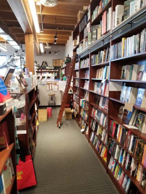 A cozy bookstore aisle lined with bookshelves, a ladder, and scattered boxes on the floor.