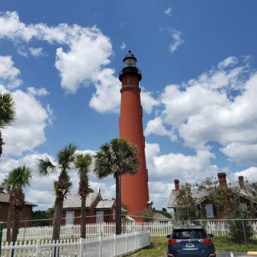 A tall red lighthouse surrounded by palm trees and historic buildings under a blue sky with fluffy white clouds.