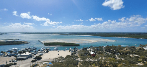 Aerial view of a coastal landscape with sandy banks, blue waters, and boats, under a bright, partly cloudy sky.