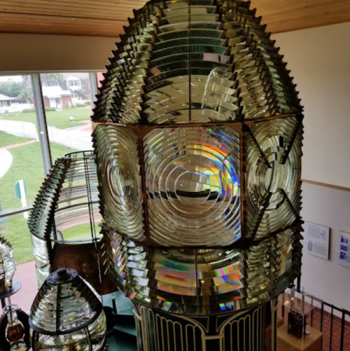 A close-up view of a large, intricate lighthouse lens with colorful glass panels, set in a bright, airy space.