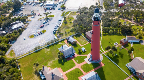 Aerial view of a red lighthouse surrounded by green grass and buildings, with a parking area in the background.
