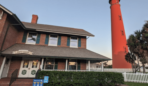 A brick building with a gift shop sign next to a tall red lighthouse, surrounded by greenery and a white fence.