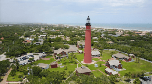 Aerial view of a red lighthouse surrounded by green trees and buildings, near a beach and ocean in the background.