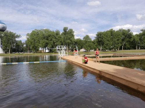 Two lifeguards on a wooden dock by a swimming area, with trees and a blue sky in the background.