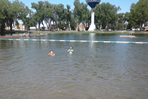 Two children swim in a calm lake surrounded by trees, with a water tower visible in the background.
