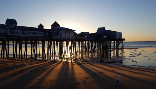 Sunset over a pier with buildings on stilts, casting long shadows on the sandy beach. Seagulls are visible in the foreground.