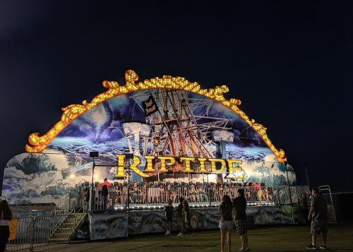 A brightly lit carnival ride named "Riptide" with a ship design, set against a dark evening sky. People are waiting in line.