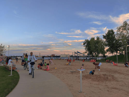 A sandy beach area with children playing, adults relaxing, and a cyclist passing by under a colorful sunset sky.