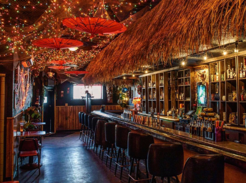 A vibrant tiki bar with a thatched roof, colorful lights, and a long bar lined with stools.