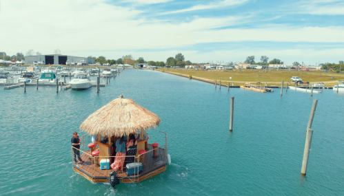 A floating tiki bar with people enjoying drinks, surrounded by boats in a calm blue waterway under a clear sky.