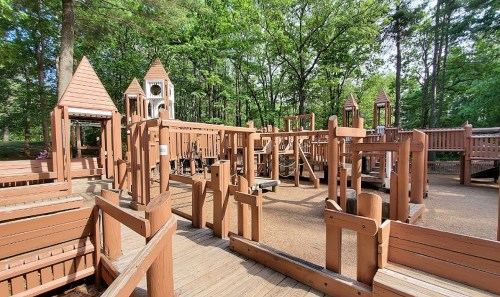 A wooden playground with various structures surrounded by trees in a sunny park setting.