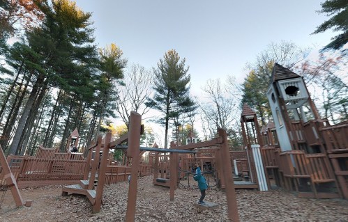 A playground surrounded by trees, featuring wooden structures and a child playing on a swing.