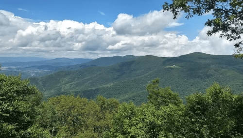 A panoramic view of lush green mountains under a blue sky with fluffy white clouds.