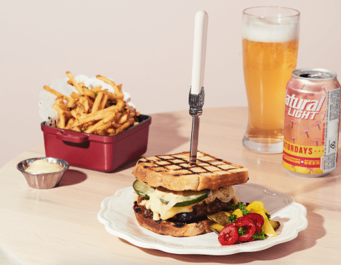 A gourmet sandwich with pickles and sauce, served with fries, a beer, and a can of soda on a wooden table.