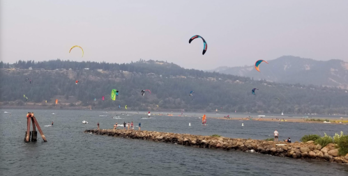 A scenic view of a lake with colorful kites and windsurfers, surrounded by mountains and a hazy sky.