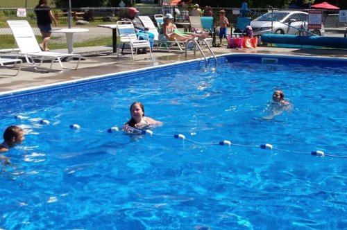 Two people swim in a bright blue pool, with lounge chairs and a sunny background.