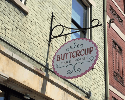 Sign for "Buttercup Cake House" hanging from a building, featuring decorative lettering on a pastel background.