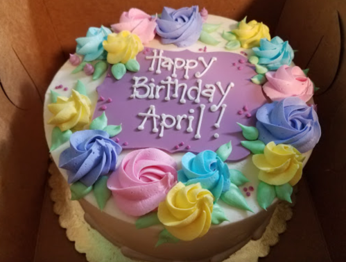 A round birthday cake with colorful rose decorations and the message "Happy Birthday April!" on a purple background.