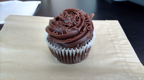 A chocolate cupcake with swirled frosting and chocolate sprinkles, sitting on a brown paper surface.