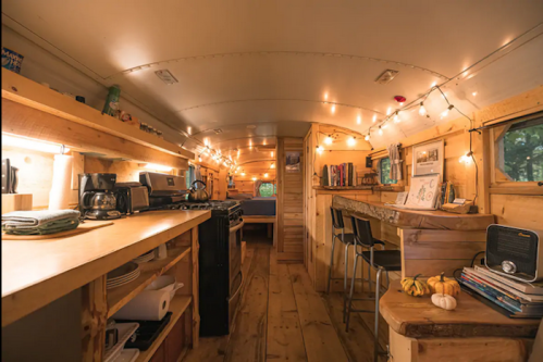 Cozy interior of a converted bus with wooden shelves, string lights, kitchen area, and a small dining space.