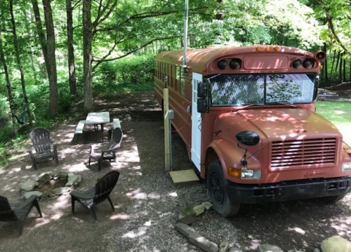 An orange school bus parked in a wooded area, surrounded by chairs and a fire pit.