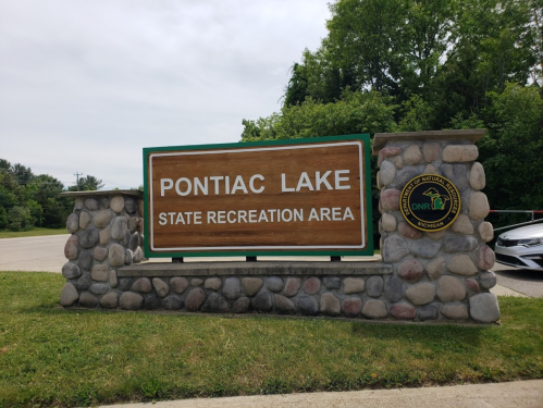 Sign for Pontiac Lake State Recreation Area, featuring a wooden board and stone base, surrounded by greenery.
