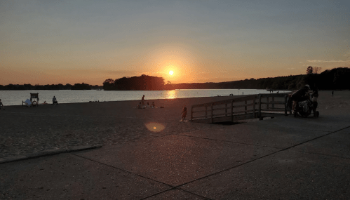 Sunset over a beach with people enjoying the sand and water, and a wooden walkway leading to the shore.