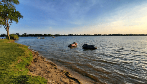 Two jet skis are anchored near the shore of a calm lake, with trees and a clear sky in the background.