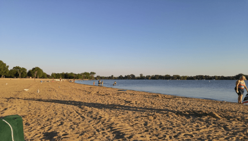 A sandy beach with people enjoying the water and trees lining the shore under a clear blue sky.