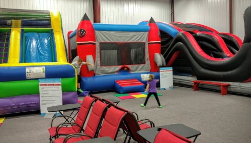 A colorful indoor play area with inflatable slides and bounce houses, and empty red chairs in the foreground.