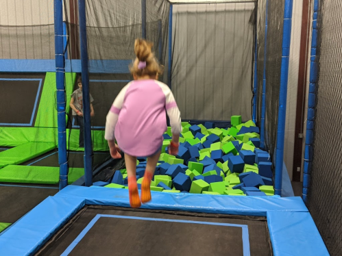 A child in a pink shirt jumps into a foam pit filled with green and blue blocks at a trampoline park.