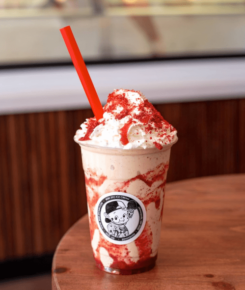 A milkshake topped with whipped cream and red drizzle, served in a clear cup with a red straw and a logo.