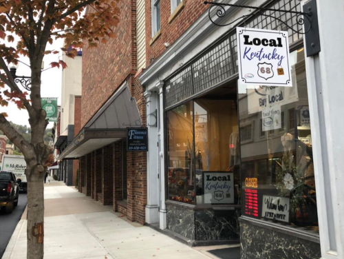 A storefront with a "Local Kentucky" sign, featuring a window display and nearby trees along a brick sidewalk.