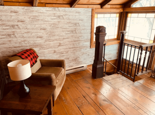 Cozy interior with a wooden staircase, a chair with a plaid blanket, and a lamp on a small table. Natural light from windows.