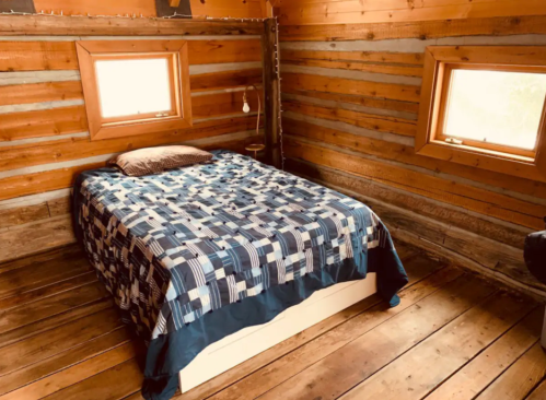 Cozy cabin bedroom with a wooden interior, featuring a bed with a blue patterned blanket and two windows.