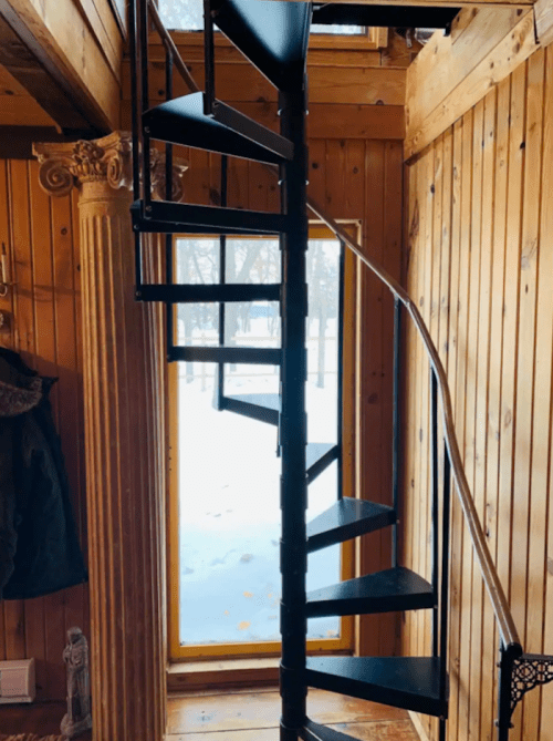 A spiral staircase leads up from a wooden interior, with a snowy landscape visible through a large window.