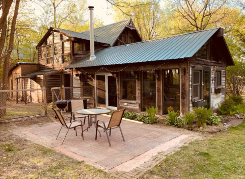 A rustic house with a metal roof, surrounded by trees, featuring a patio with a table and chairs.
