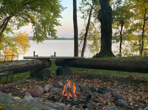 A cozy campfire surrounded by logs and stones, with a serene lake and trees in the background at sunset.