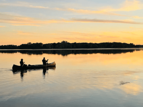 Two people canoeing on a calm lake at sunset, with vibrant colors reflecting on the water's surface.
