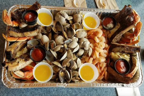 A seafood platter featuring shrimp, clams, crab, ribs, and dipping sauces in a large aluminum tray.