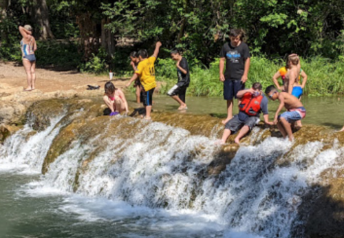 Children playing near a waterfall, some splashing in the water while others stand on the rocks. A woman watches nearby.