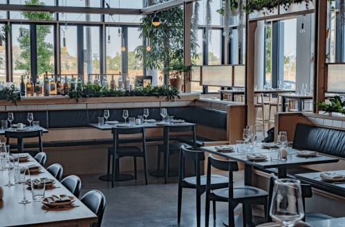 Modern restaurant interior with large windows, wooden tables, and greenery, creating a bright and inviting atmosphere.