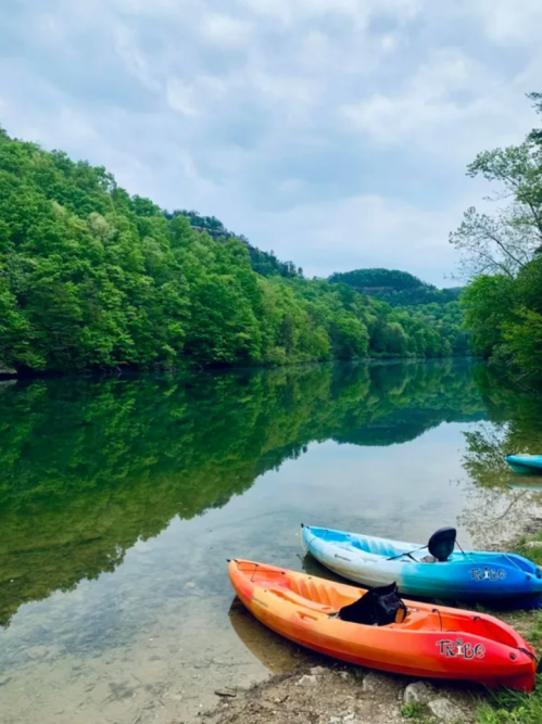 Two kayaks, one orange and one blue, are parked on a sandy shore beside a calm, reflective river surrounded by lush greenery.