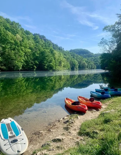 Colorful kayaks lined up on a sandy shore beside a calm, reflective river surrounded by lush green trees.