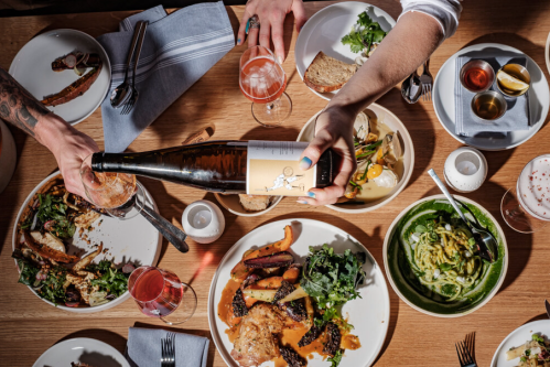 A table filled with various dishes, drinks, and hands pouring wine, creating a vibrant dining experience.