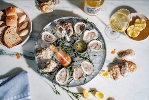 A platter of oysters on ice, garnished with lemon, sauce, and accompanied by bread and drinks on a marble surface.