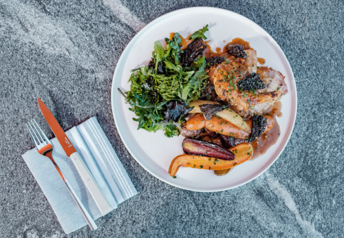 A plate of roasted chicken with vegetables, greens, and morel mushrooms, accompanied by a knife and fork on a napkin.