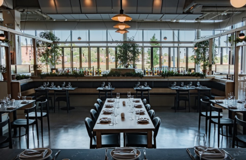 A modern restaurant interior with large windows, wooden tables set for dining, and greenery in the background.