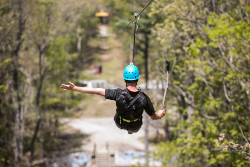 A person in a blue helmet zip-lining through a forested area, with a path visible below.