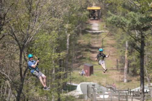 Two people ziplining through a forested area, wearing helmets and harnesses, with a zipline platform in the background.